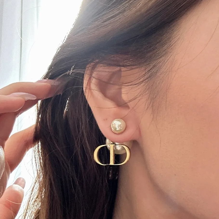 A close-up of a person's ear wearing a gold double-C hoop earring with a pearl stud, with dark brown hair and a hand adjusting a strand of hair in the foreground.