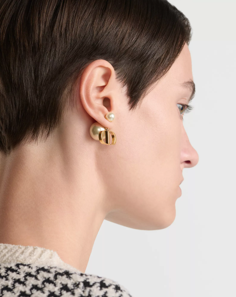 A close-up side profile of a model with short dark hair wearing a gold hoop earring with a small pearl stud.