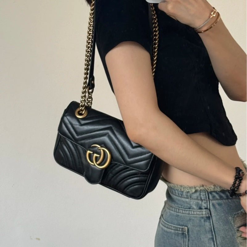 Black handbag with gold chain strap held by a person wearing a black top and blue jeans.