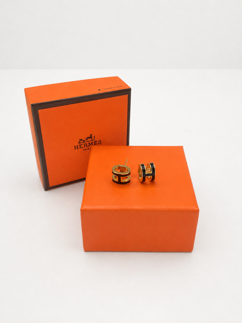 A pair of orange and black hoop earrings displayed on top of an orange presentation box, with the matching orange box standing behind them on a white background.
