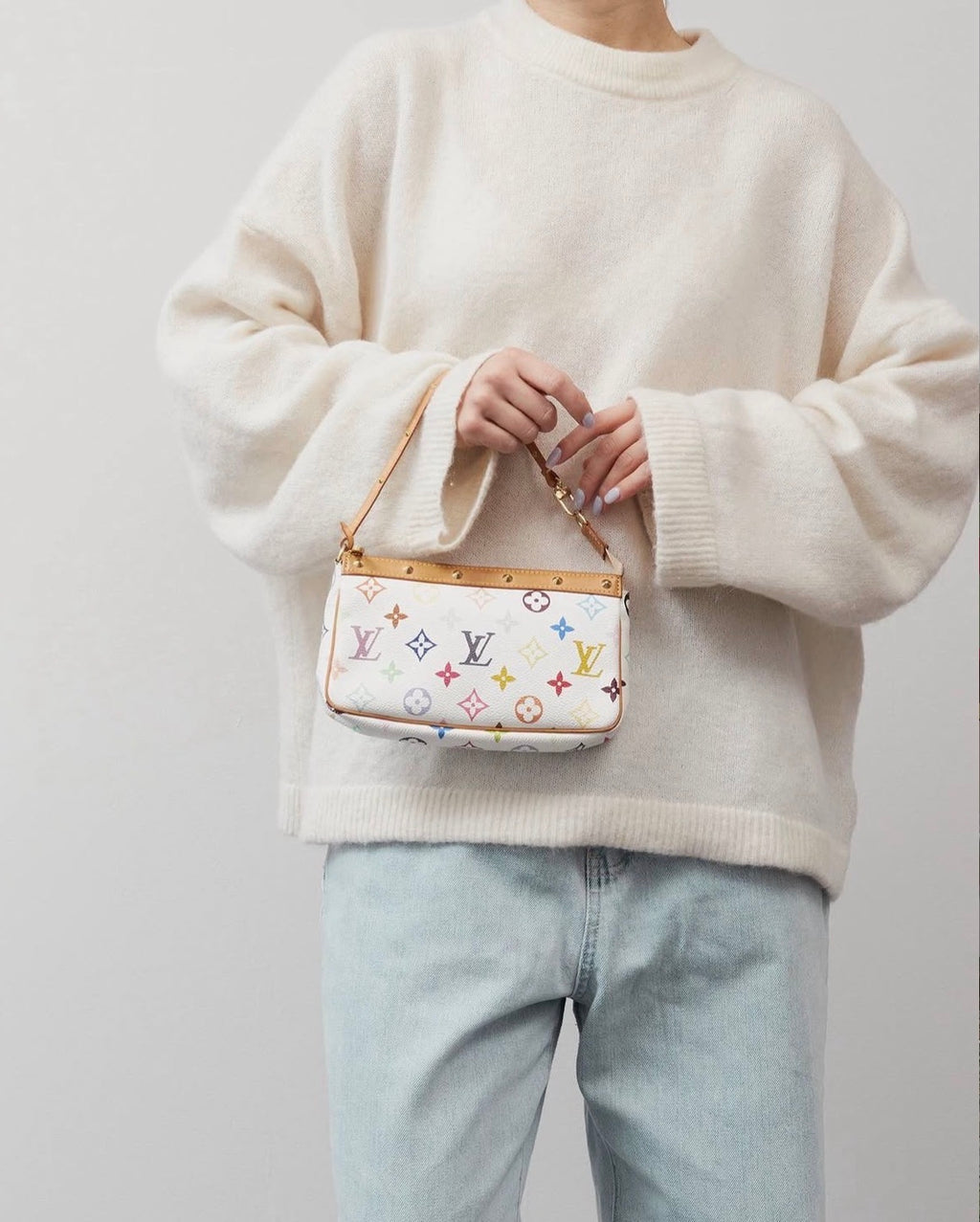 A person wearing an oversized cream knit sweater and light blue jeans holds a small multicolor bag. The bag is white with a multicolor  pattern and gold tone hardware.