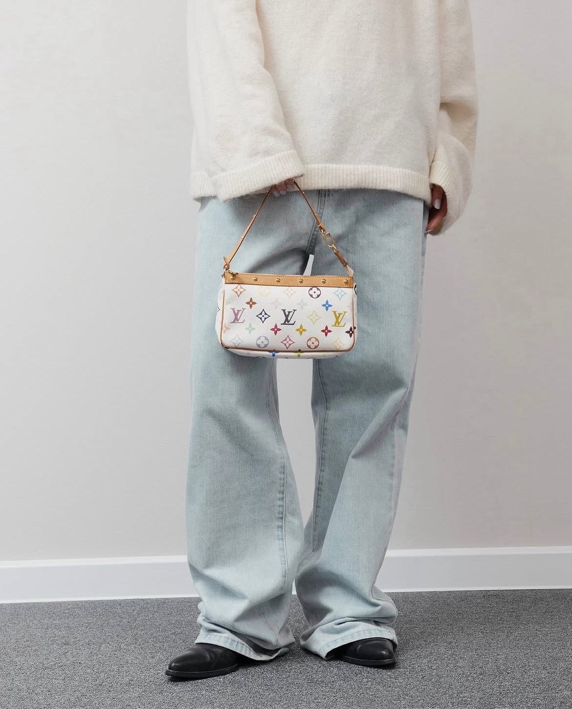 A person wearing a cream knit sweater, light-wash wide-leg jeans, and black shoes holds a small bag. The bag is white with a multicolor pattern and gold-tone hardware, shown against a plain light gray wall and gray floor.